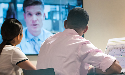 Two people in a meeting room looking at a TV that displays a man in business attire.
