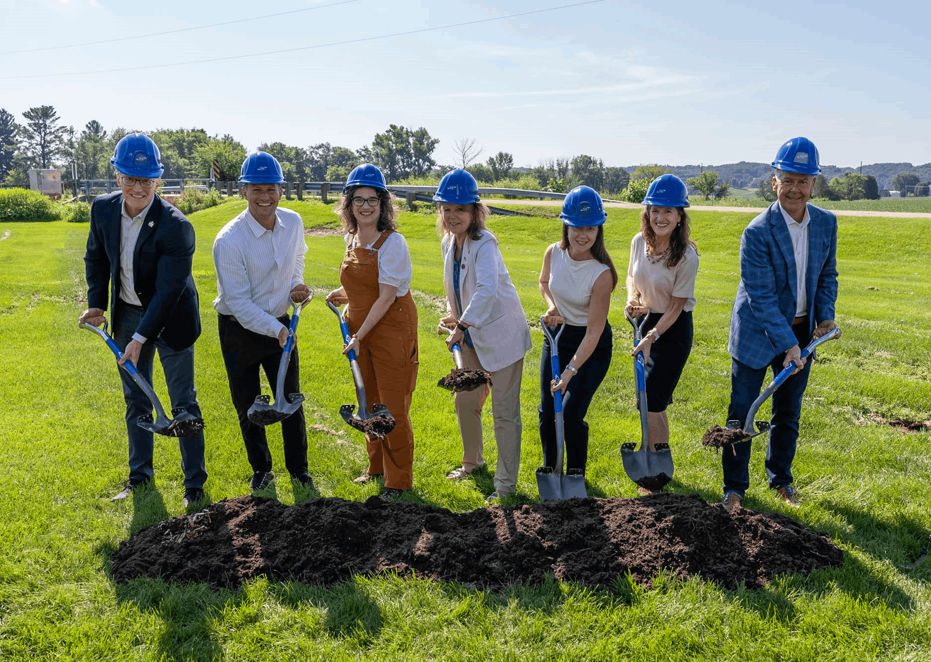 A group of seven people wearing blue construction helmets smile while holding shovels, participating in a groundbreaking ceremony on a sunny day.