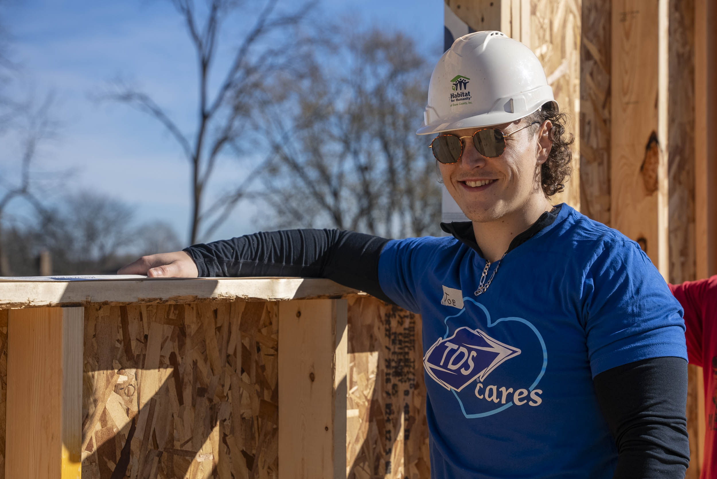 A caucasian male wearing a white hard hat, sunglasses, and a blue TDS t-shirt standing next to a construction area and smiling