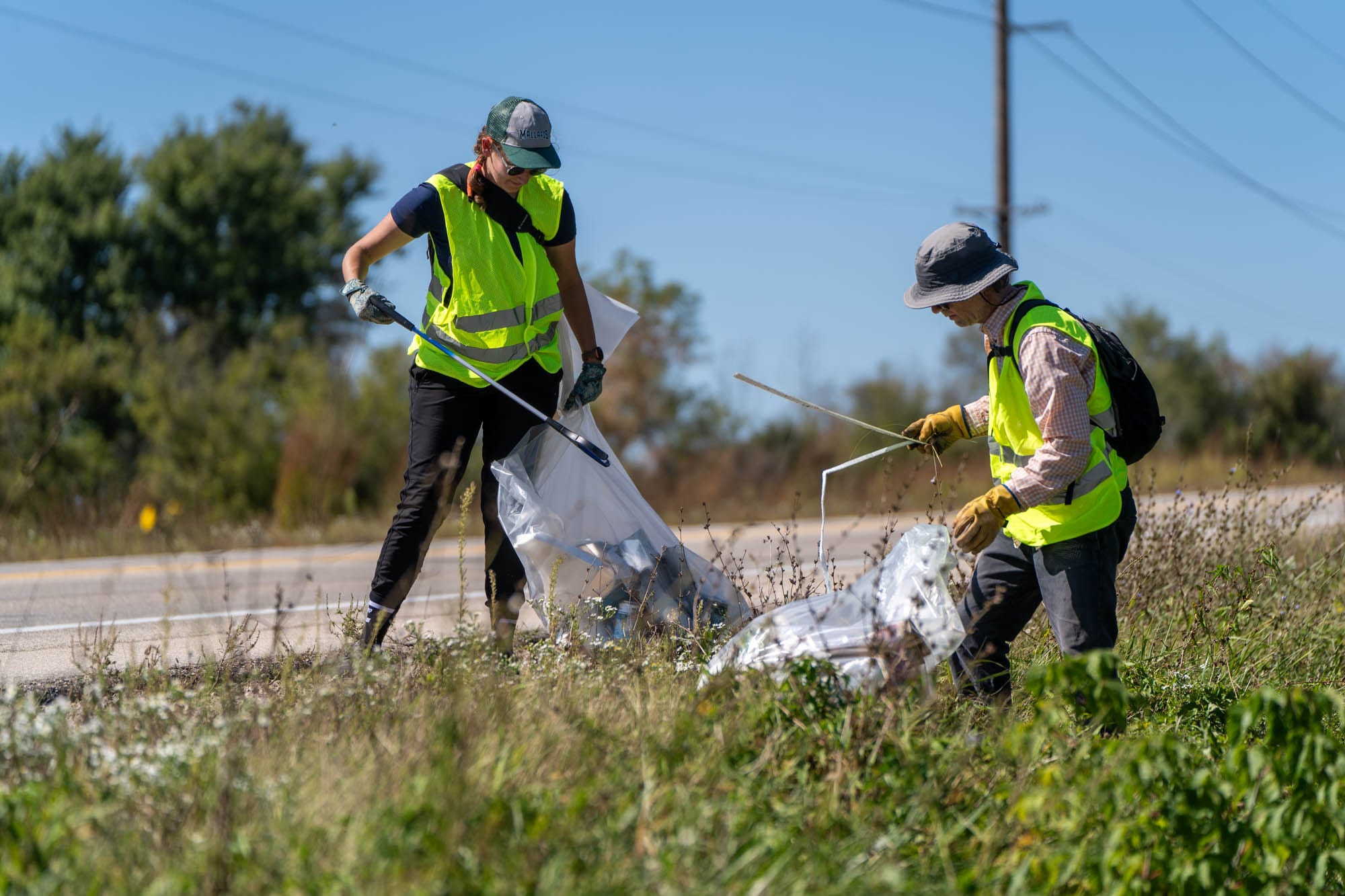 A woman and a man wearing reflective yellow vests picking up trash off of the side of a highway