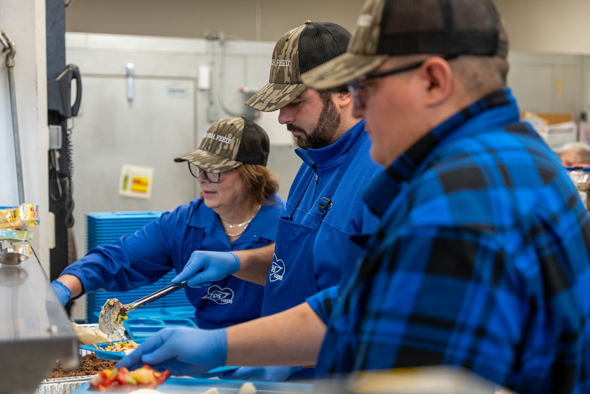 Two men and one woman wearing blue TDS aprons and camo hats in a kitchen scooping out food to give to hungry people