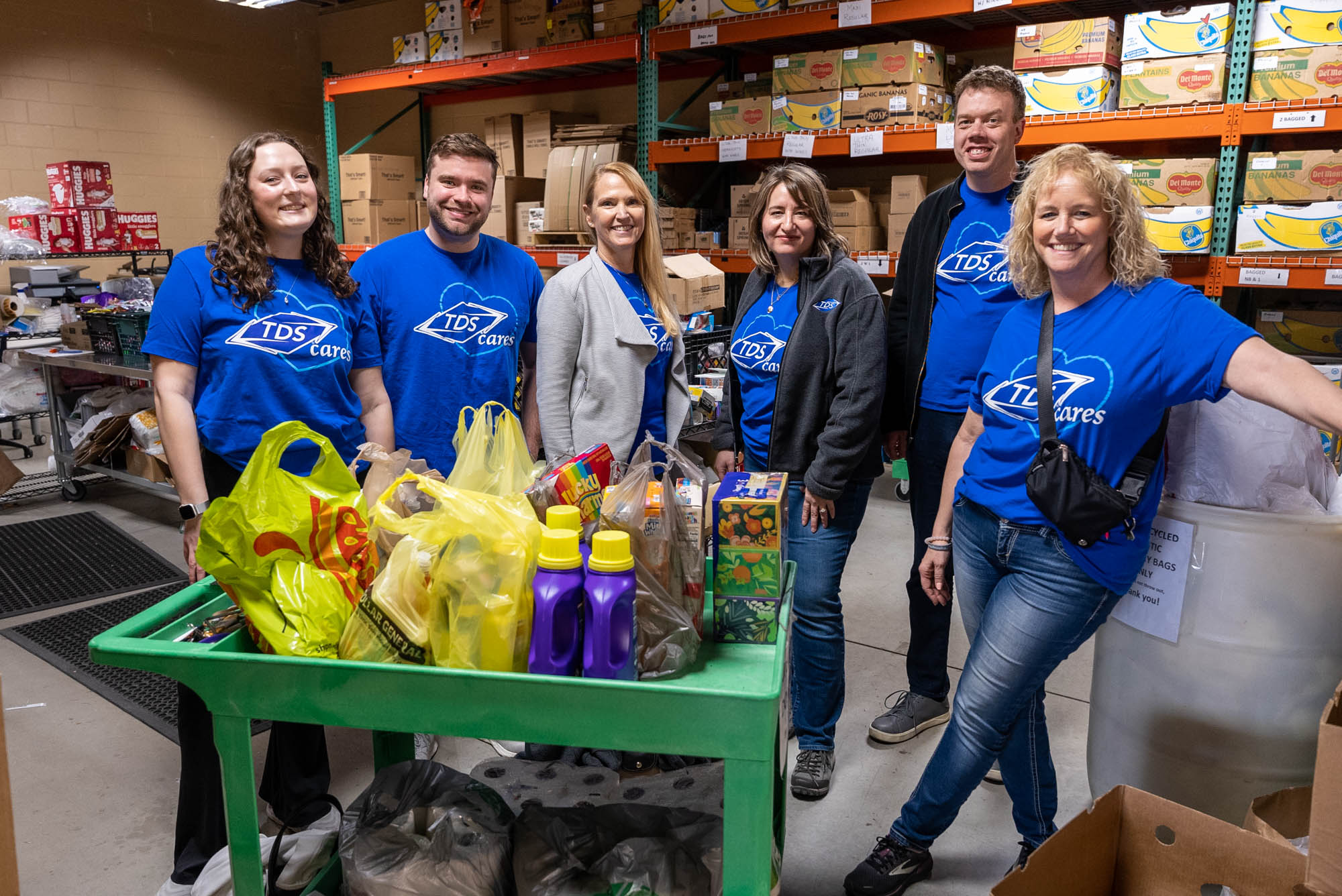 Four women and two men all wearing blue TDS t-shirts, standing in a warehouse behind a cart hold a bunch of household items like kleenex, detergent, and toilet paper