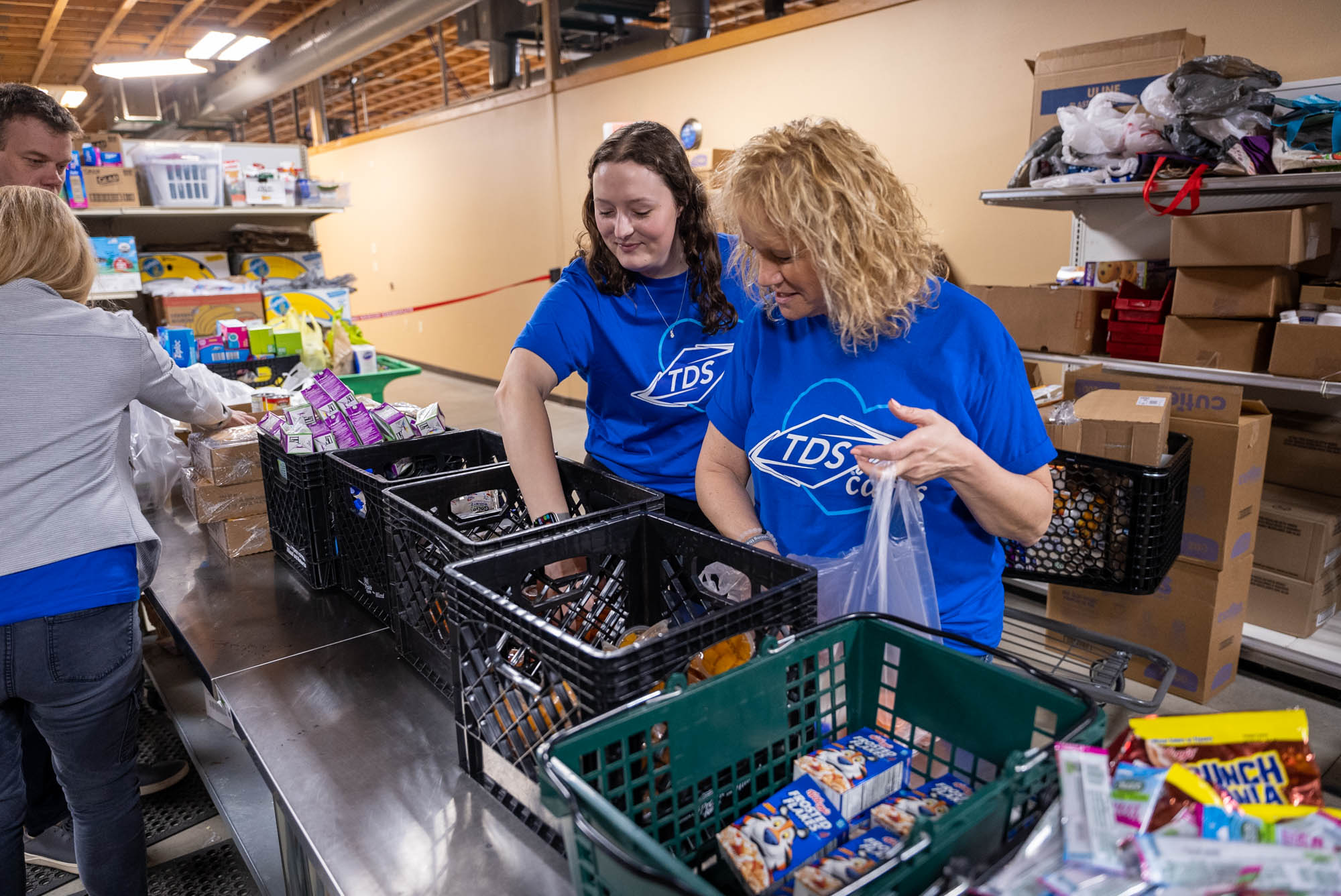 Two women wearing blue TDS t-shirts packing groceries into plastic bags from crates