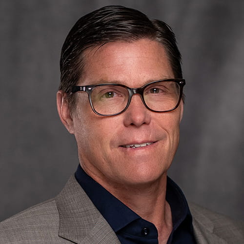 A man with short, dark hair wearing glasses and a gray blazer over a navy shirt, against a gray background. He has a neutral, professional expression.