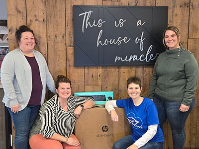 Four women smile in front of a wooden wall sign that reads 
