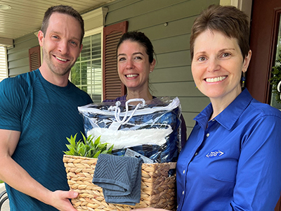 Three people stand smiling on a porch. One holds a gift basket containing towels and a small plant. The mood is cheerful and welcoming.