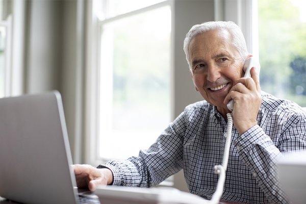 Man at home at his desk on a laptop talking on his landline phone